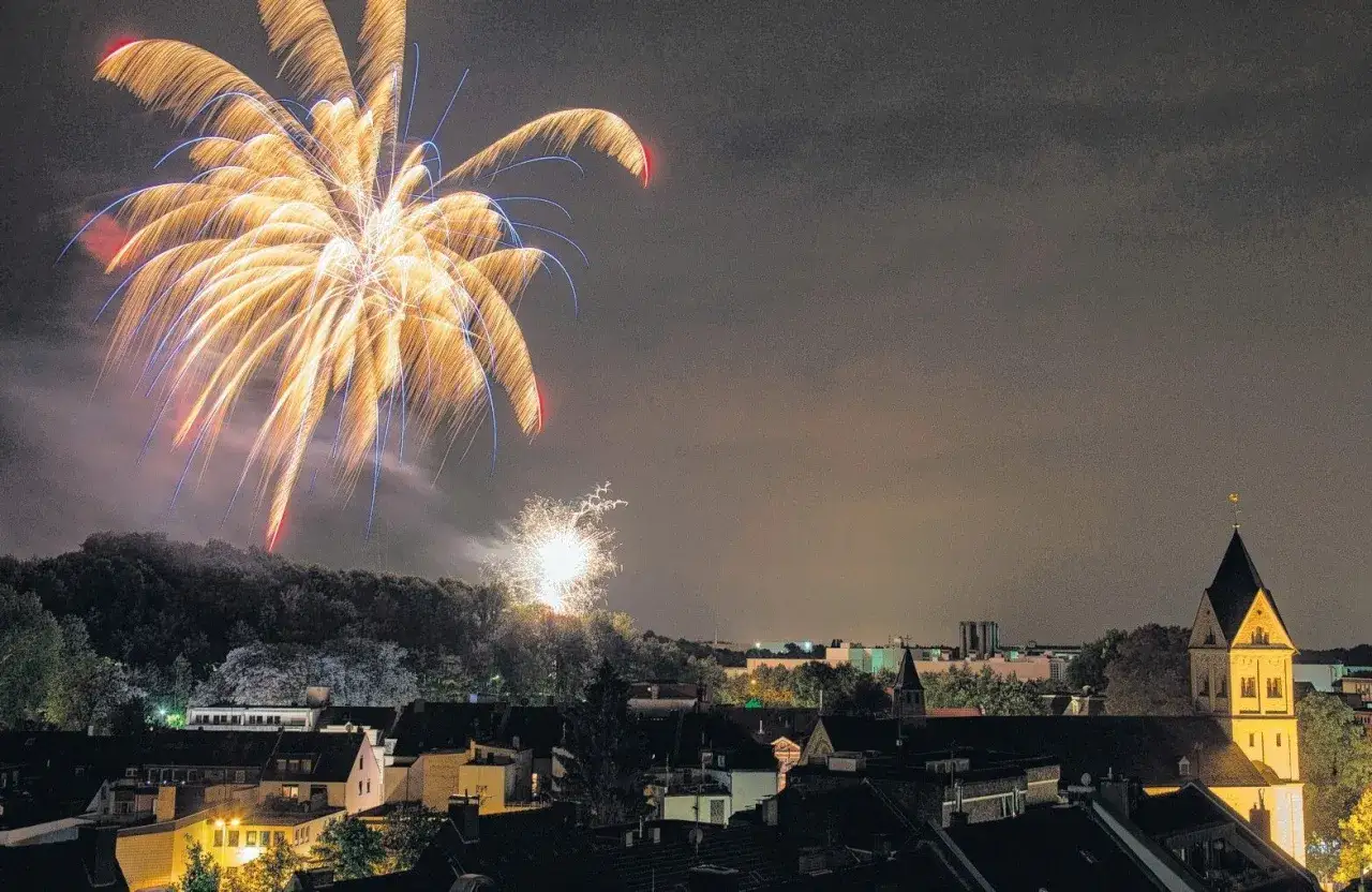 Feuerwerk &uuml;ber Bergisch Gladbach. Ein helles, goldgelbes Feuerwerk erhellt den Nachthimmel &uuml;ber der Stadt, w&auml;hrend unten die D&auml;cher der H&auml;user und ein Kirchturm zu sehen sind.