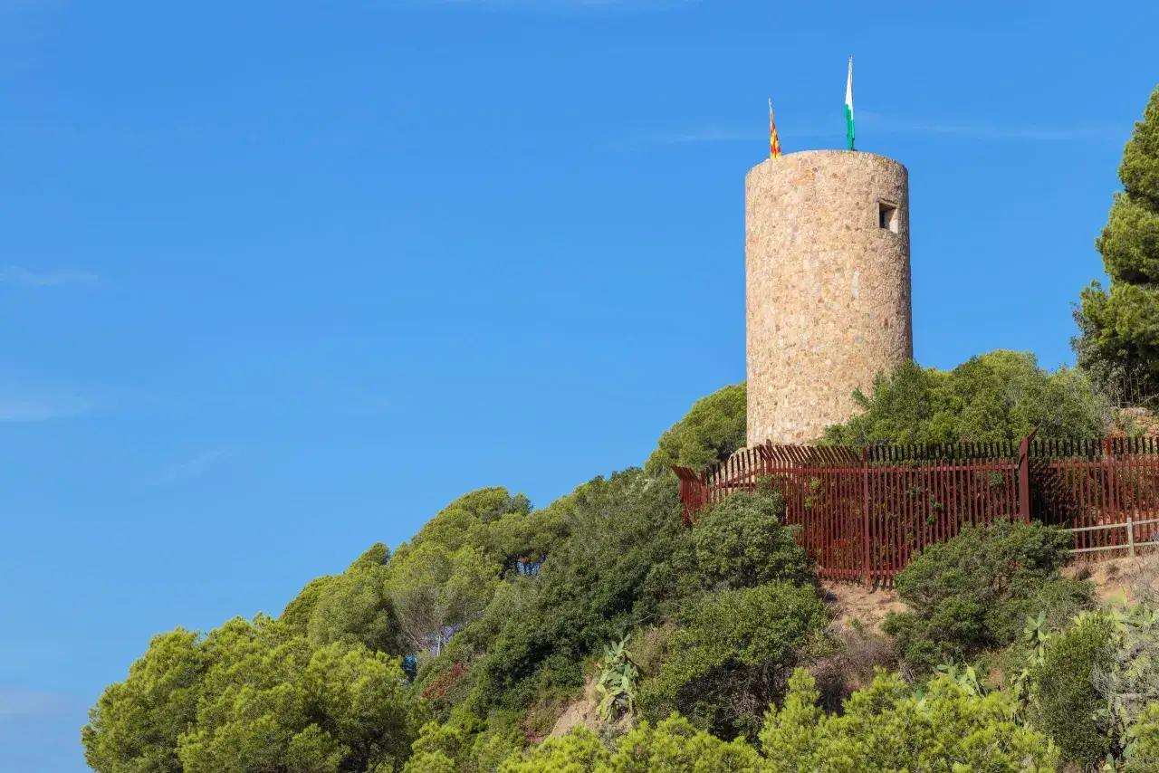 Torre de piedra con banderas catalana y andaluza en la cima, rodeada de vegetaci&oacute;n. El kasteel lloret de mar se alza vigilante.