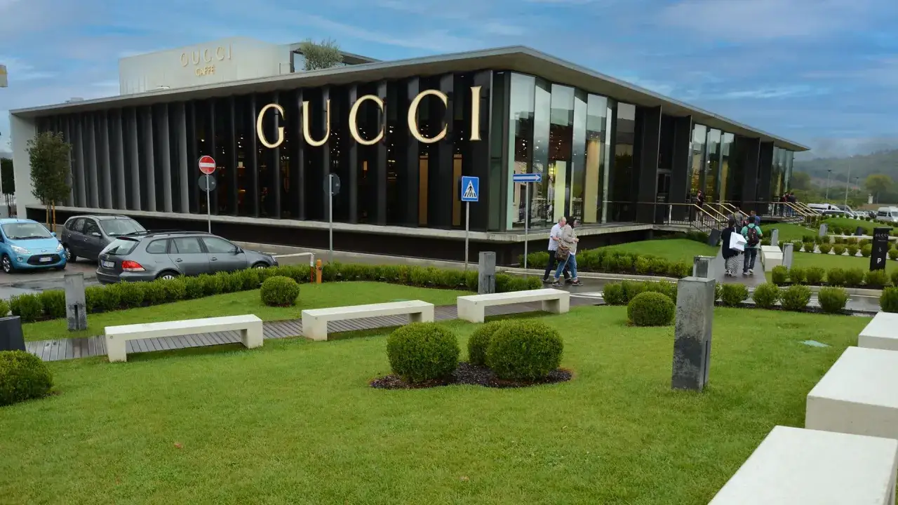 Un **outlet** en **Italie** avec le logo Gucci illuminé. Des clients se promènent sur la pelouse devant le bâtiment moderne.