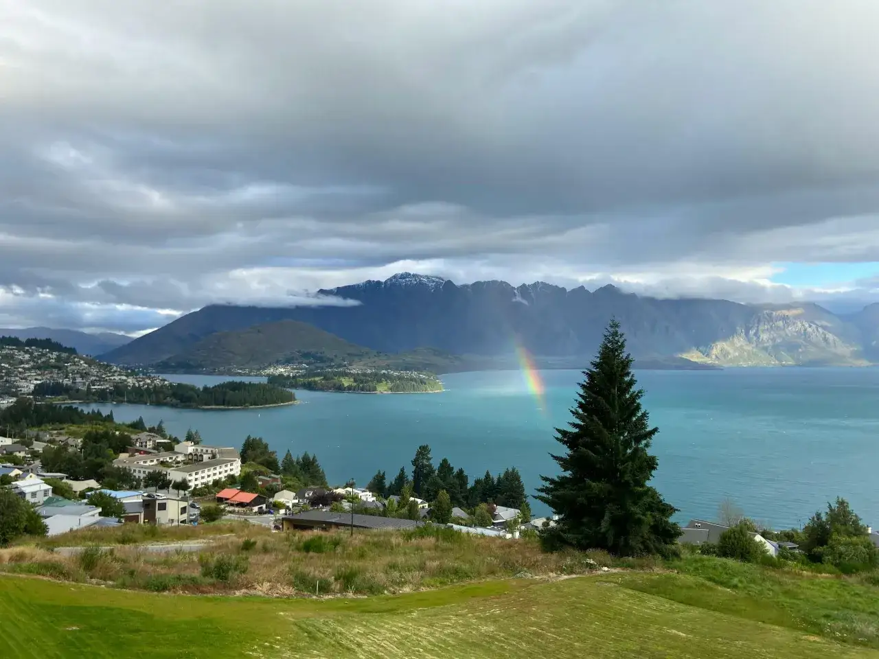 Un arcoíris aparece sobre un lago turquesa, con montañas nevadas al fondo. Un pueblo se extiende por la ladera. ¡Un paisaje que te hará pensar cuándo viajar a Nueva Zelanda!