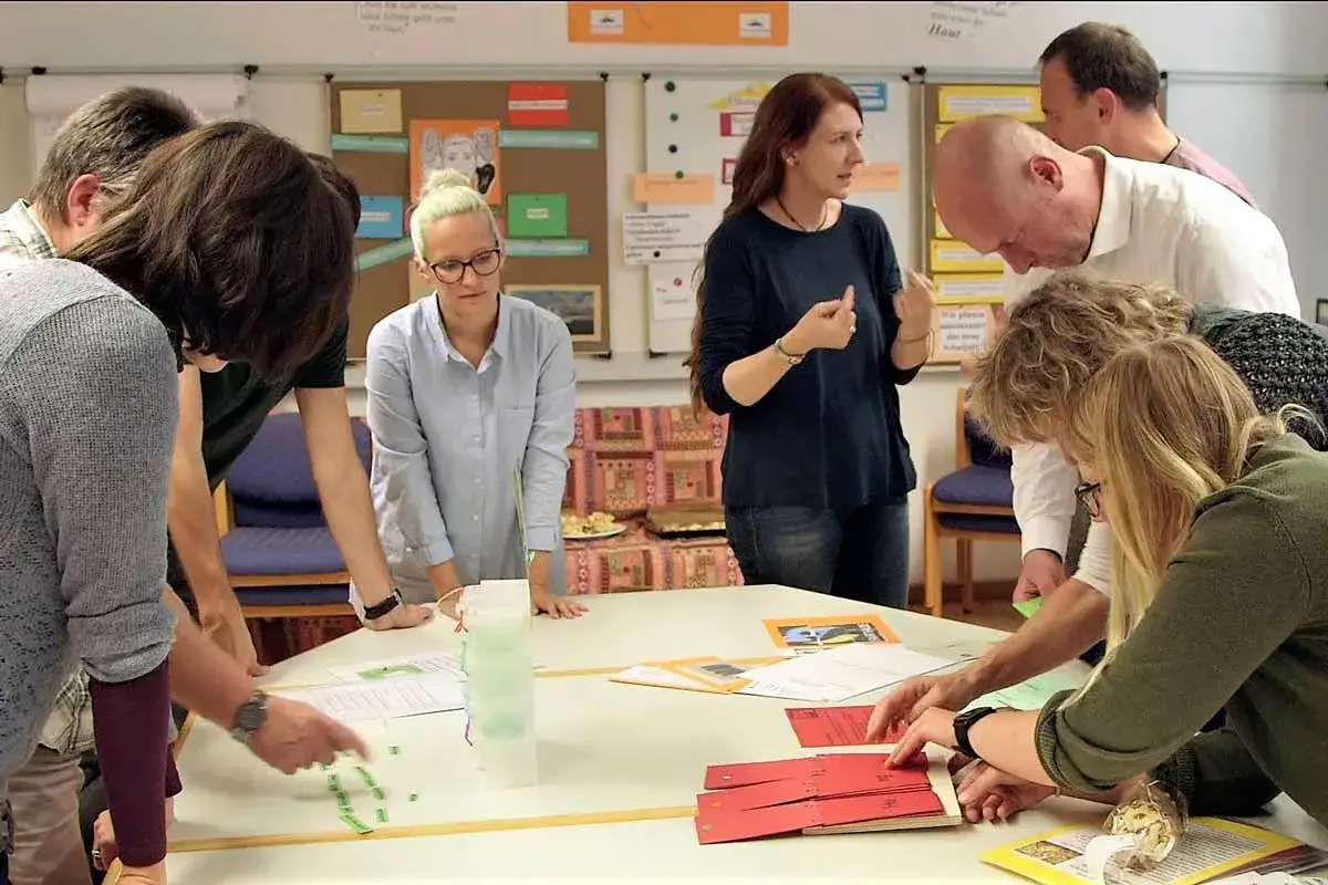 Ein Lehrerteam bespricht sich an einem Tisch, auf dem Unterlagen und kleine gr&uuml;ne K&auml;rtchen liegen.