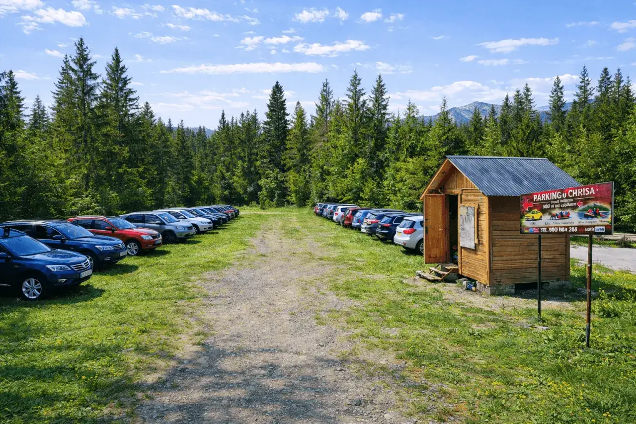 Parking przed wejściem do Doliny Chochołowskiej. Samochody zaparkowane wzdłuż leśnej drogi, obok drewniana budka z tablicą informacyjną.