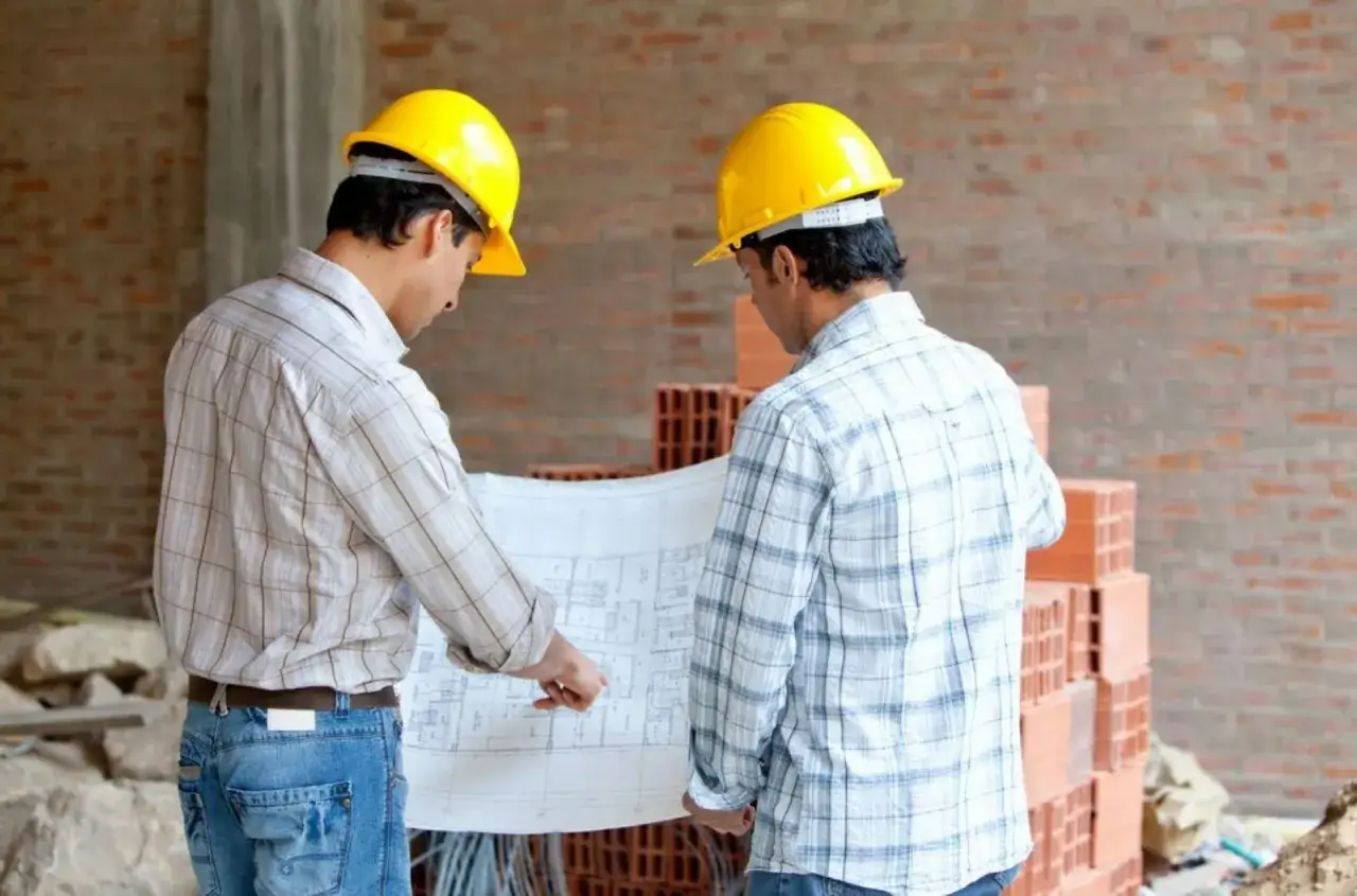 Dos ingenieros con cascos amarillos revisan planos en una obra. La direcci&oacute;n de obra avanza seg&uacute;n lo planeado.