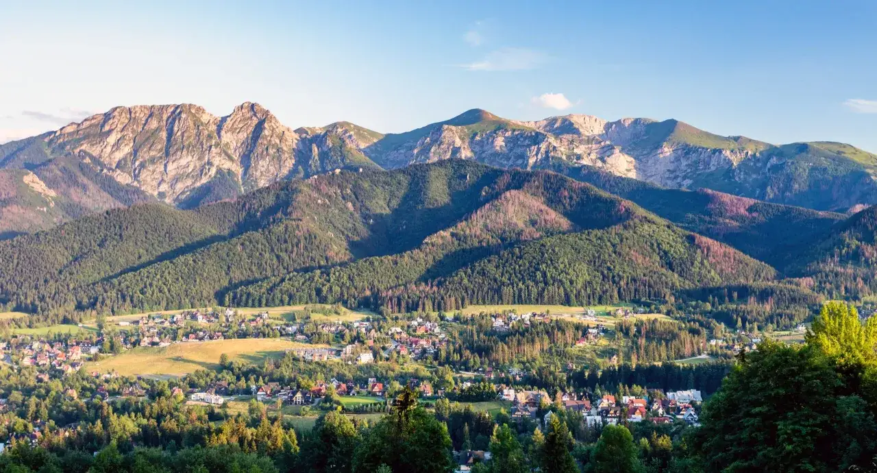 Widok na Tatry z Zakopanem u podn&oacute;ża. G&oacute;ry, lasy i zabudowania tworzą malowniczy krajobraz, gdzie przełom białki jest widoczny w oddali.