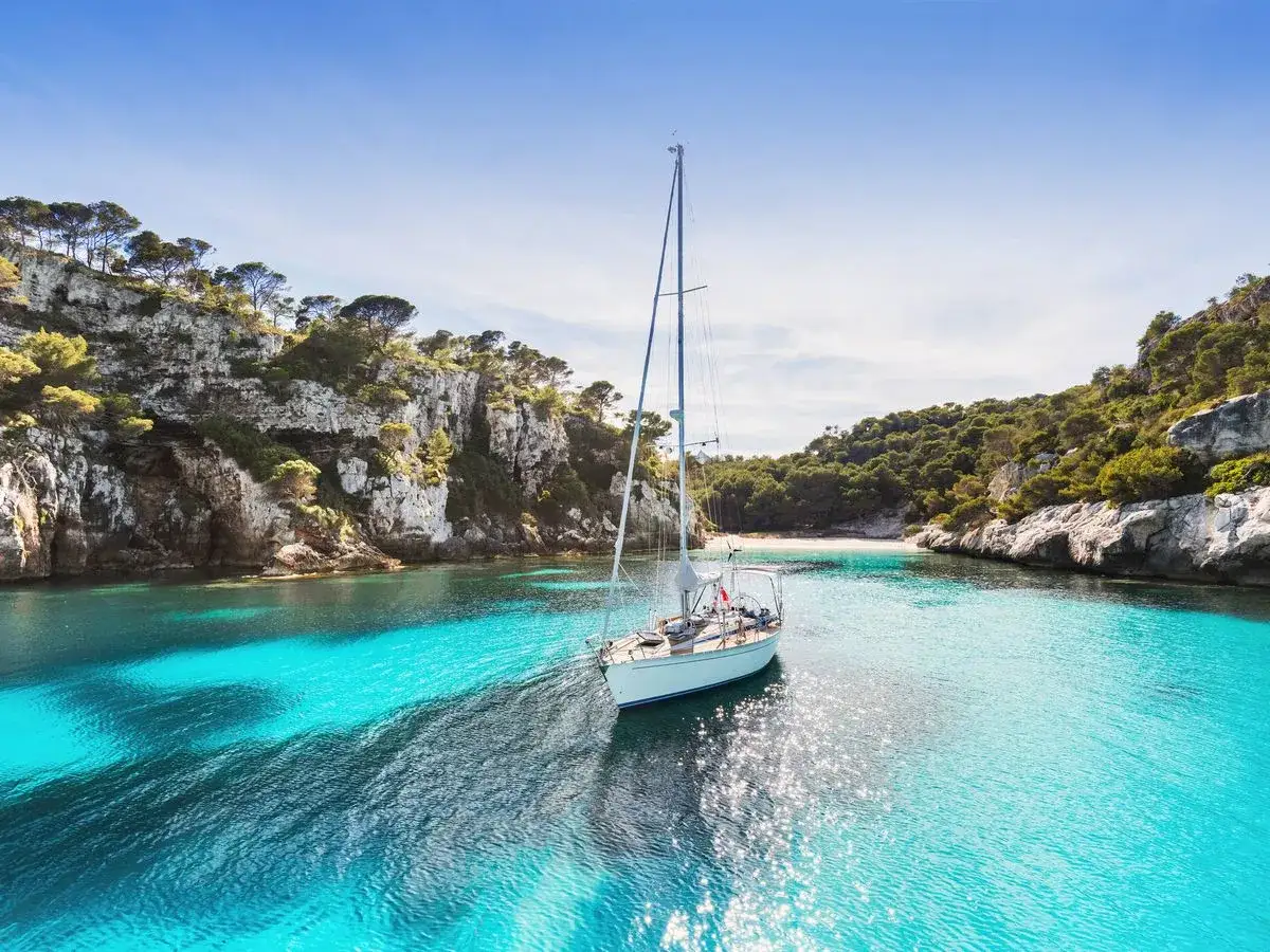 Velero navega en aguas turquesas, rodeado de acantilados y vegetación. Un sueño para las vacaciones agosto 2026.