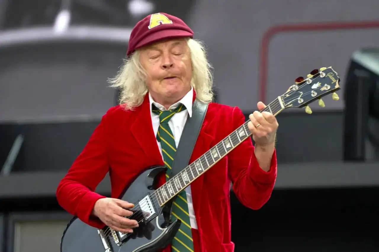 Angus Young z AC/DC gra na gitarze, ubrany w czerwony garnitur i czapkę. Jego charakterystyczne utwory rozbrzmiewają na koncercie.