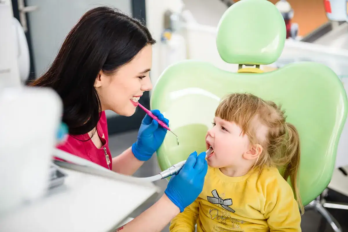 Uśmiechnięta dentystka wykonuje lapisowanie zębów u małej dziewczynki, która siedzi na zielonym fotelu.