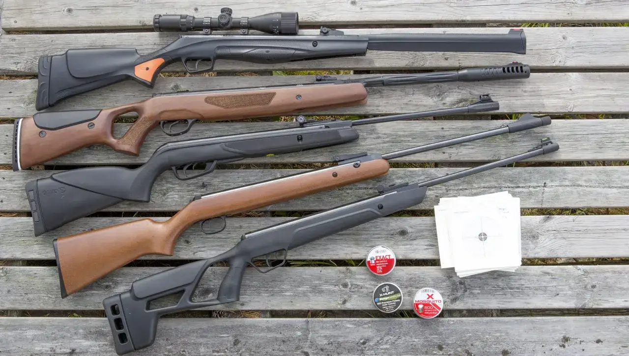 En samling kraftiga luftvapen, ammunition och måltavlor ligger utspridda på en träbänk.