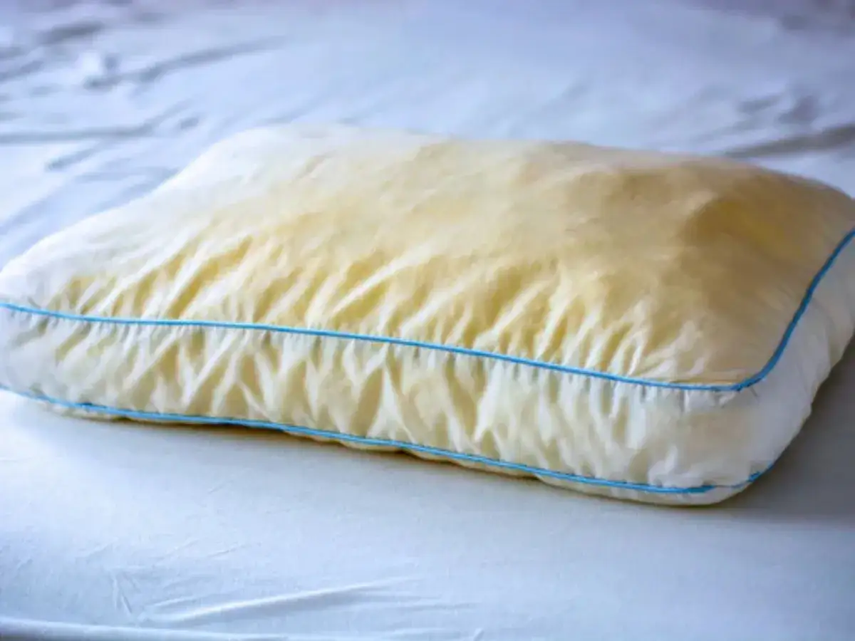 Ein vergilbtes Kissen mit blauen N&auml;hten, das auf einem wei&szlig;en Bett liegt. Gelbe Flecken deuten auf m&ouml;glichen Schimmel hin.
