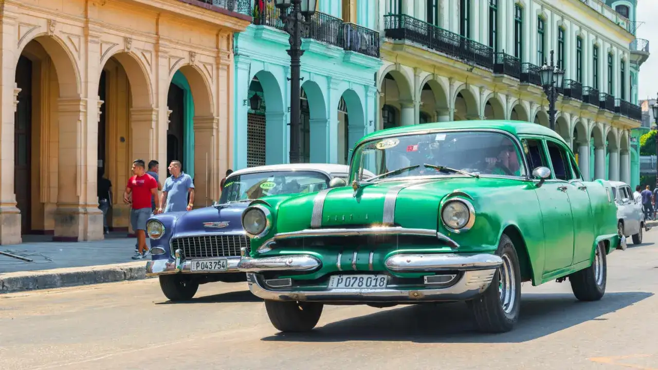 Gr&uuml;ne und blaue Oldtimer fahren durch eine Stadt mit bunten Geb&auml;uden. Ein Hauch von Nostalgie, wenn man diese Klassiker importieren w&uuml;rde.