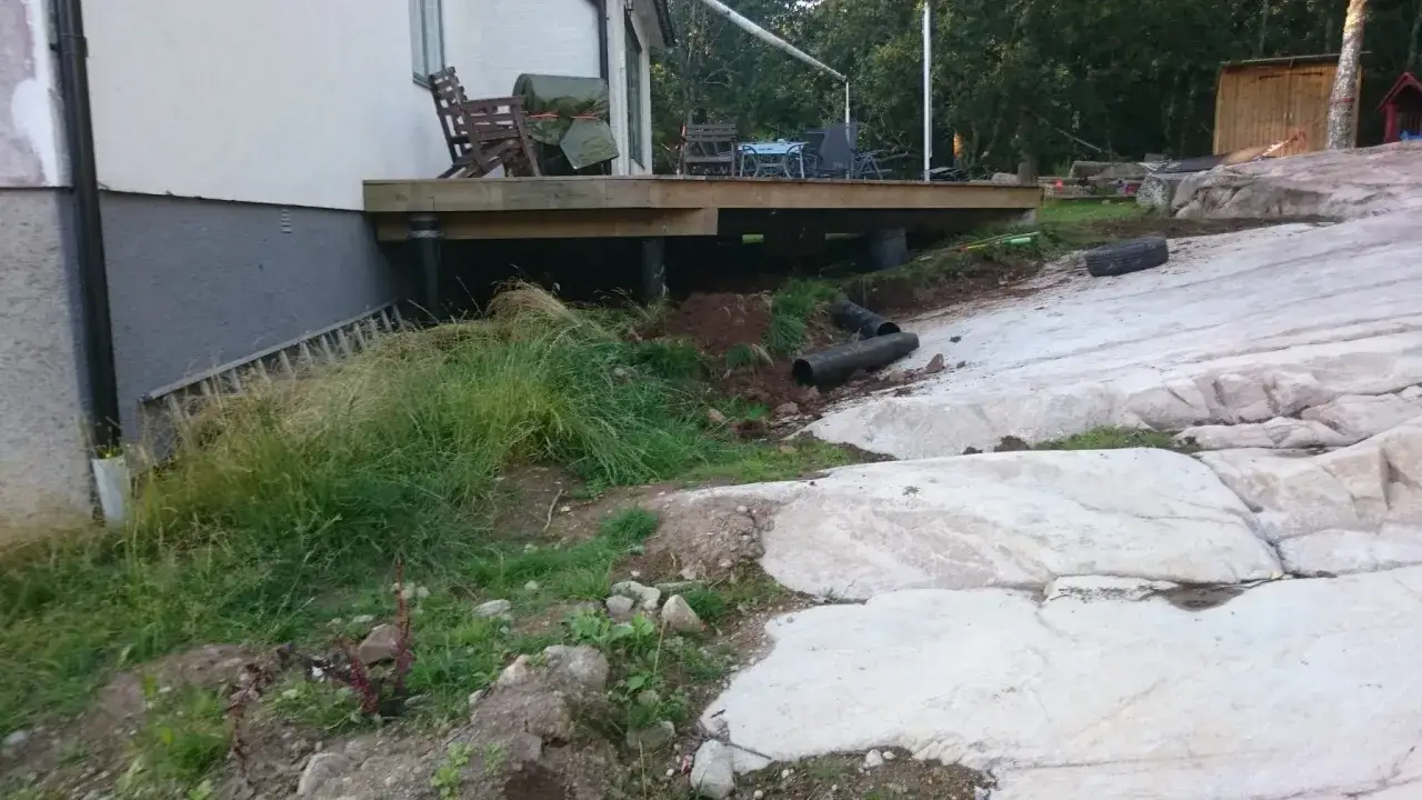 Nybyggd altan med synliga stödben. Marken runt huset är kuperad med stora stenblock, och det ser ut som att man har behövt borra i berg för att få till grunden.