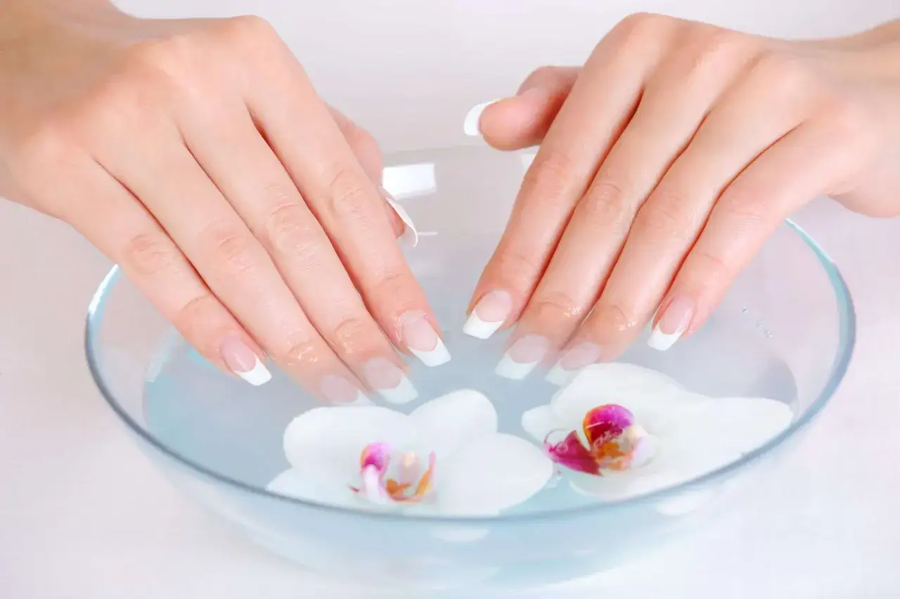 Mains soignées trempant dans un bol d'eau avec des orchidées. Une étape pour savoir comment faire pousser ses ongles.