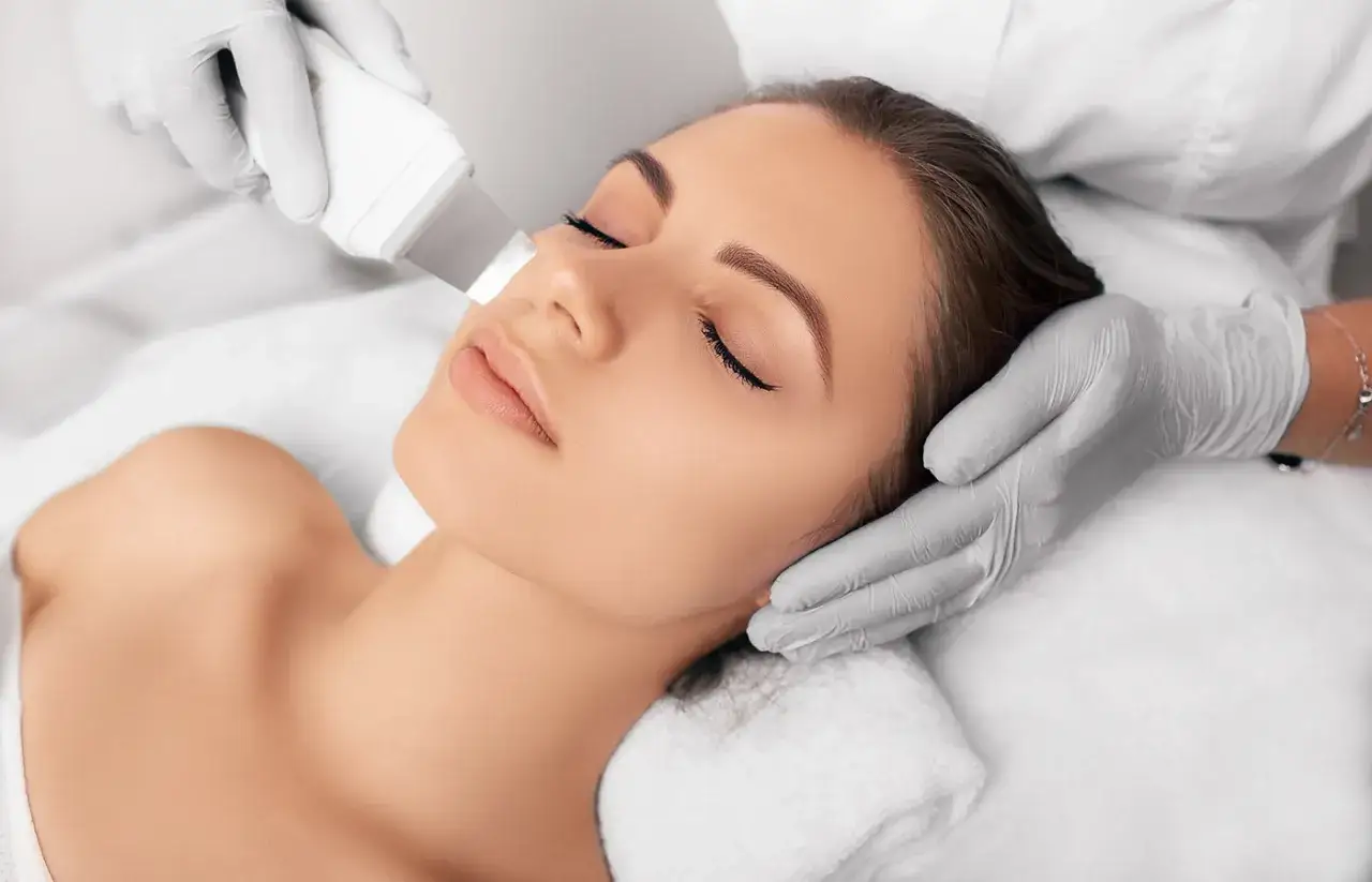 Una donna riceve un trattamento di pulizia del viso con ultrasuoni. Mani guantate tengono lo strumento che emette onde sonore per una pelle radiosa.