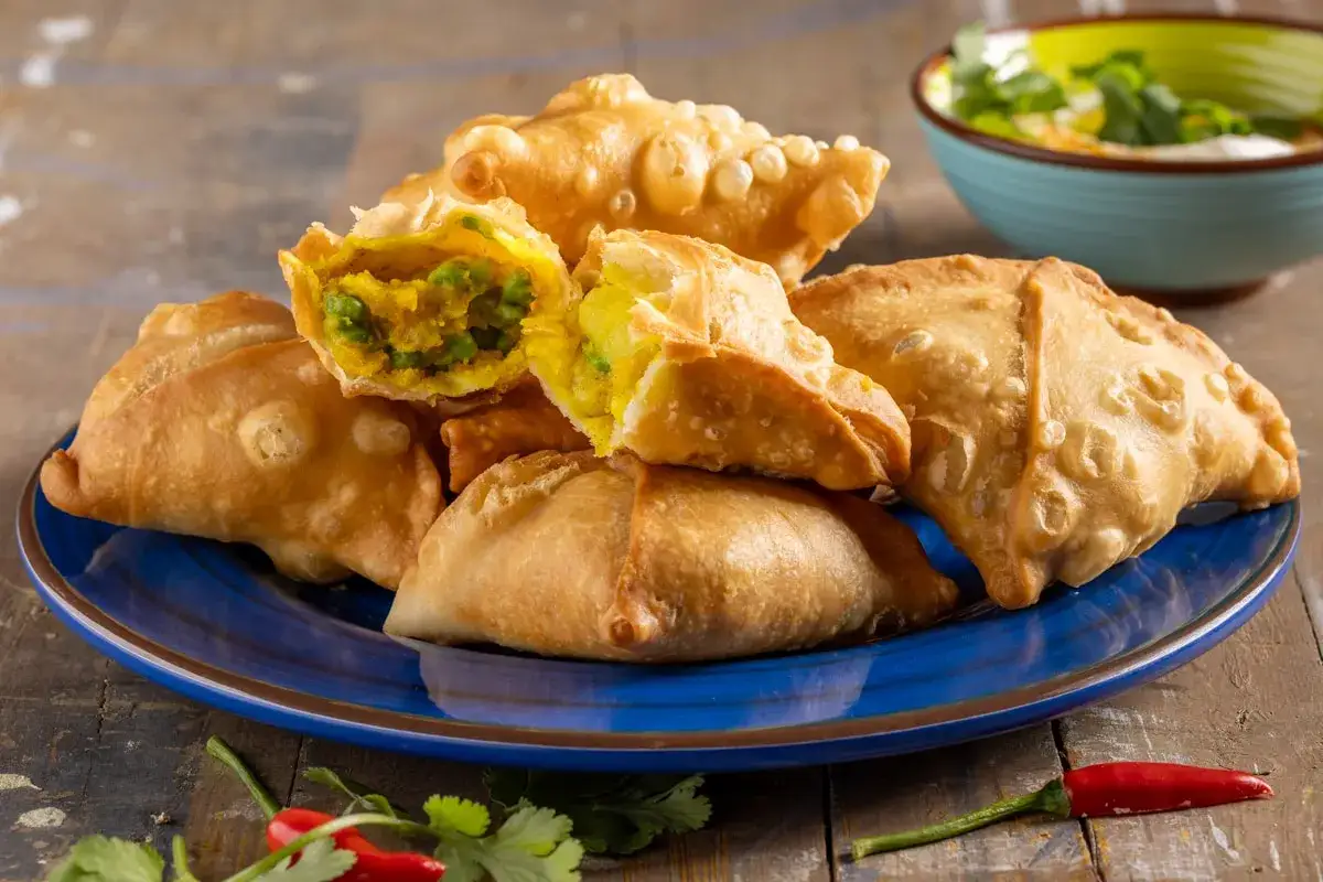 Samosa al forno dorati e croccanti, ripieni di verdure speziate, serviti su un piatto blu con peperoncino e coriandolo.