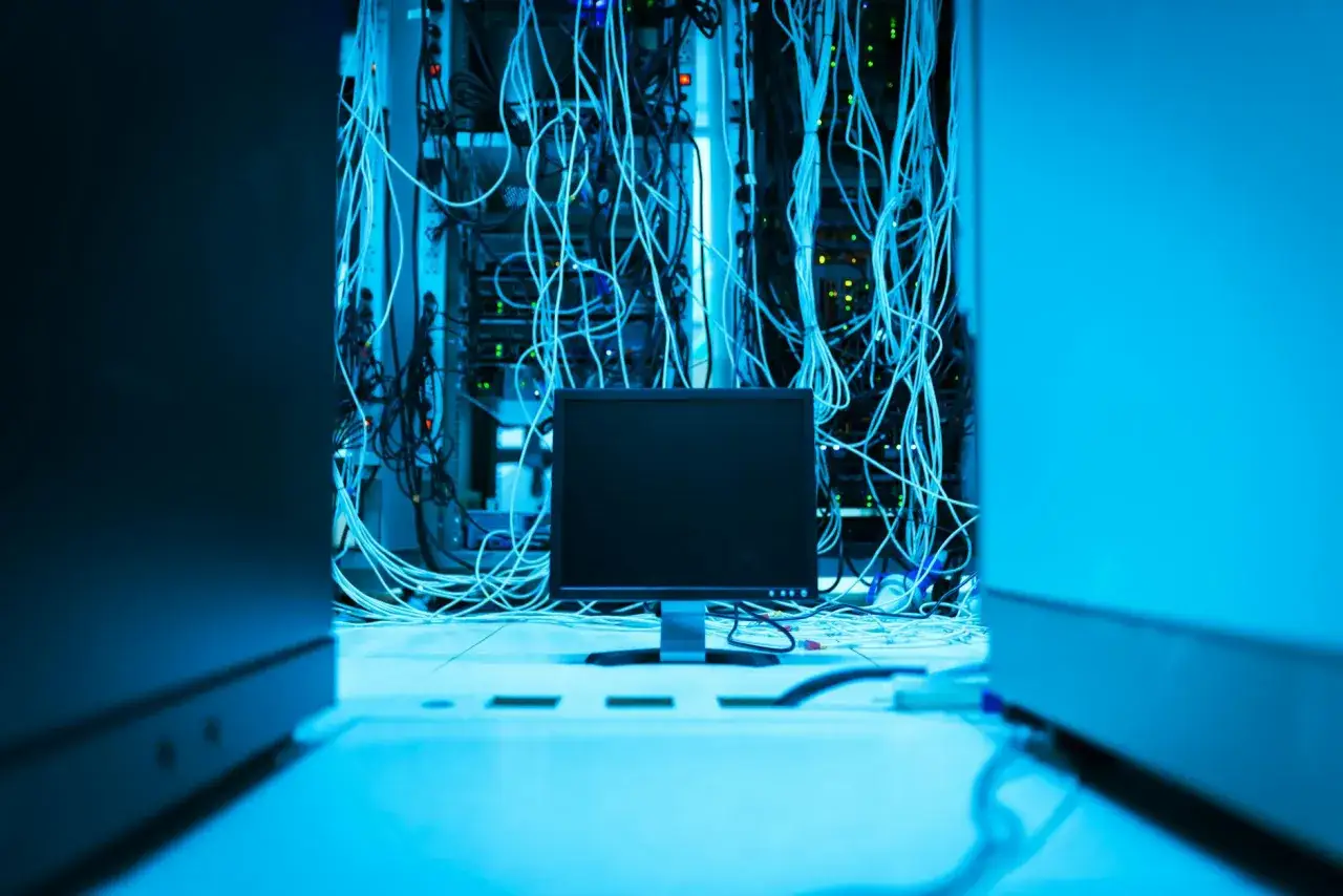 Monitor przed serwerownią z plątaniną kabli. Trwa budowa serwera, a technicy pracują nad połączeniami.