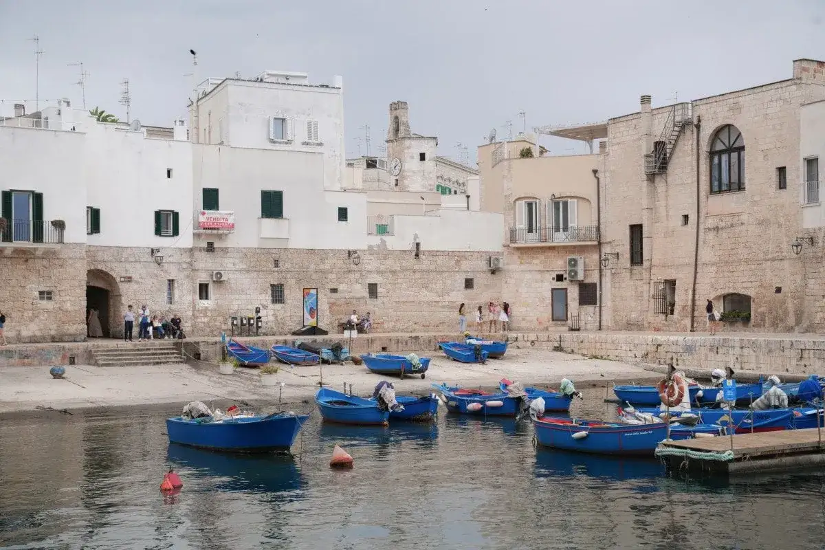 A charming harbor with blue boats, white buildings, and people enjoying the scenery. This could be the best town to stay in Puglia!