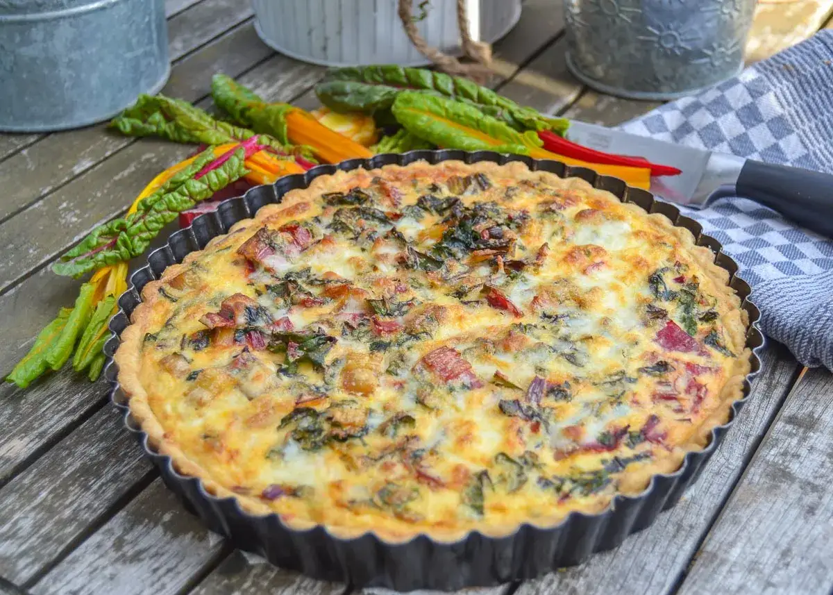 Herzhaftes Mangold Quiche Rezept, frisch gebacken und bereit zum Genie&szlig;en. Bunte Mangoldstiele liegen daneben.