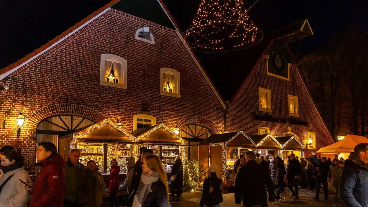 Besucher bummeln über den velen Waldweihnachtsmarkt, vorbei an festlich beleuchteten Buden und Backsteinhäusern. Ein Lichterbaum funkelt.