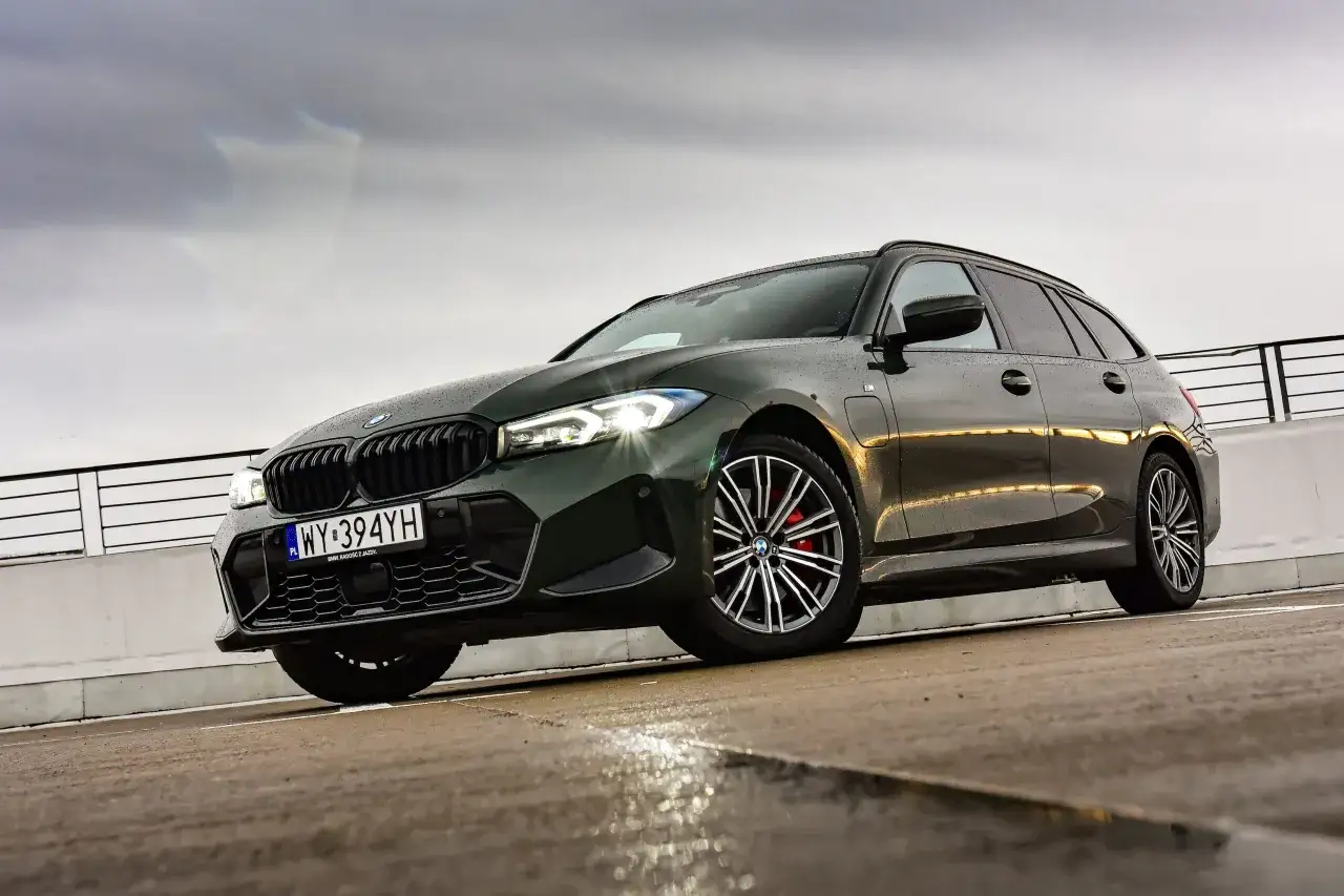 Ciemnozielone BMW 330e kombi na mokrym parkingu, z reflektorami włączonymi.