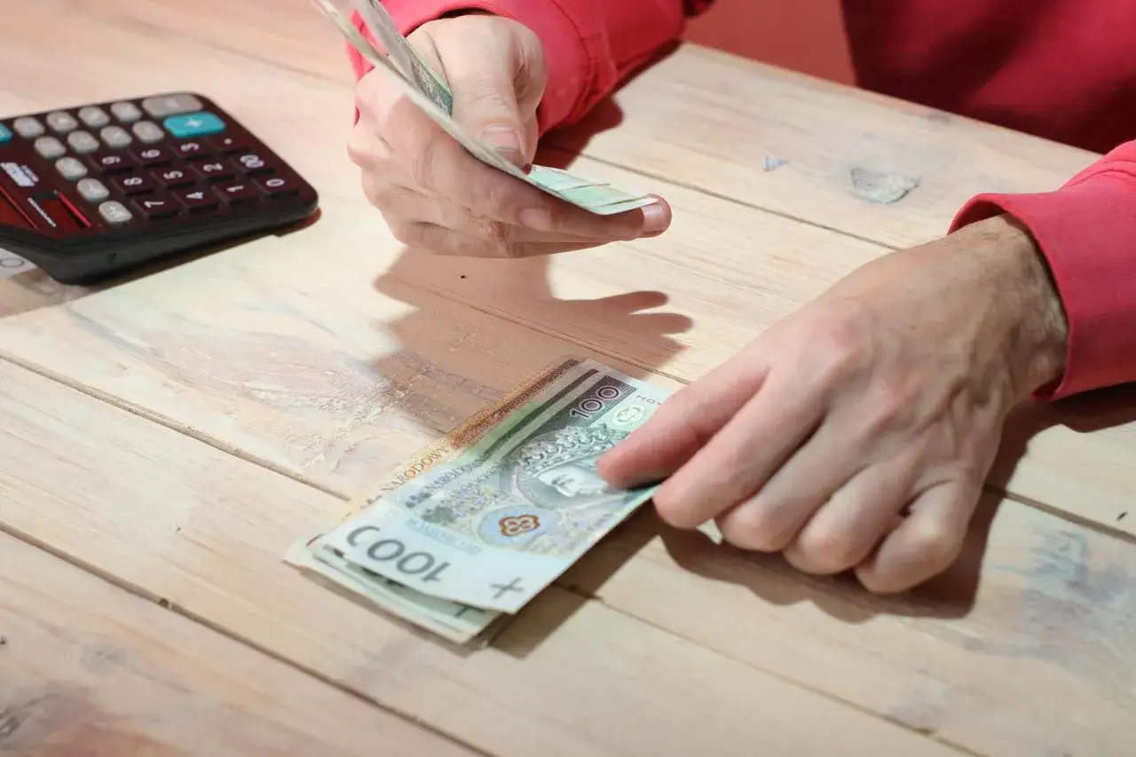 Ręce liczą pieniądze na drewnianym stole. Obok leży kalkulator. Zastanawiasz się, ile wynosi renta socjalna netto?