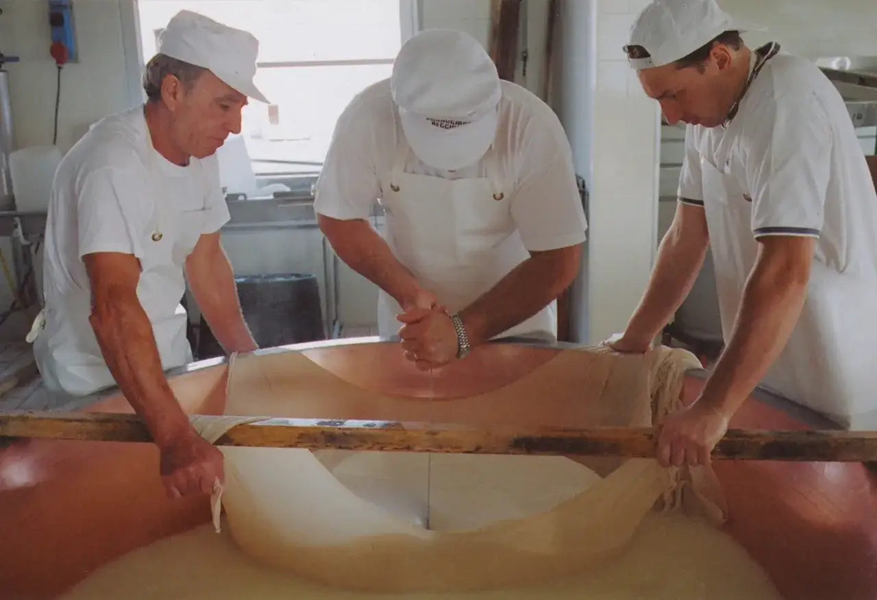 M&auml;nner in wei&szlig;en Kappen und Sch&uuml;rzen arbeiten mit einer gro&szlig;en K&auml;semasse. Sie formen den Teig, aus was Parmesan besteht, in einem Kupferkessel.