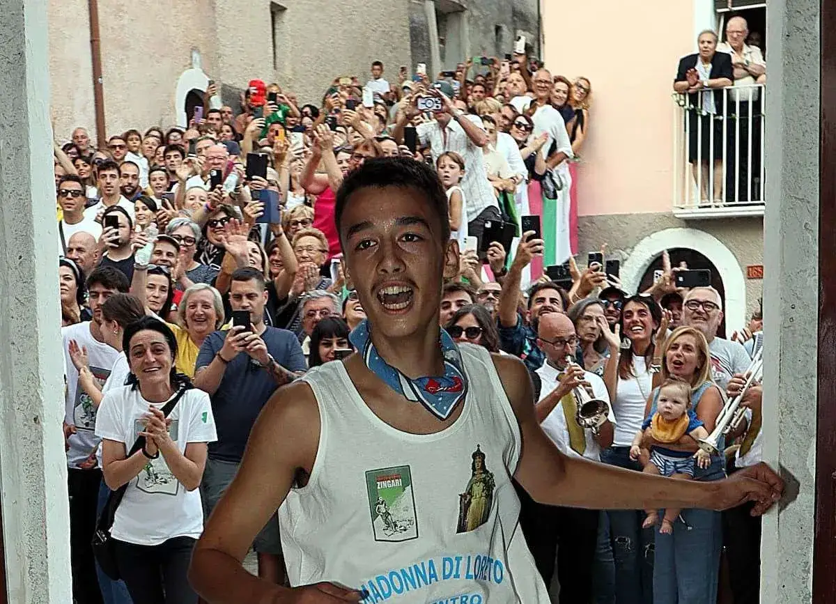Giovane festante in una processione a Pacentro, con la folla che filma e applaude. Sul suo gilet, un'immagine della Madonna di Loreto.