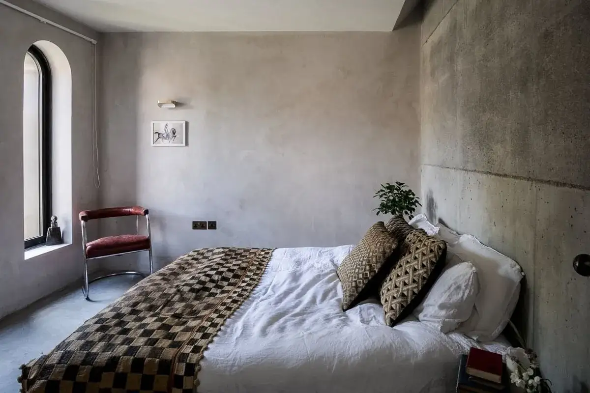 Camera da letto con pareti in intonaco argilla grezzo, letto con coperta a scacchi e cuscini decorati, sedia in pelle e finestra ad arco.