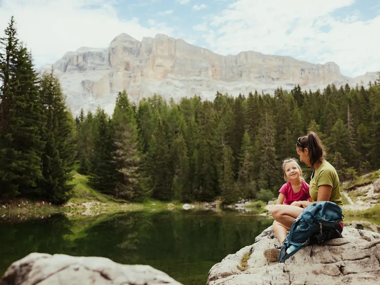 Mamma e figlia sedute su una roccia vicino a un lago alpino, con uno zaino blu. Un momento di relax con vista sulle montagne, perfetto per un 