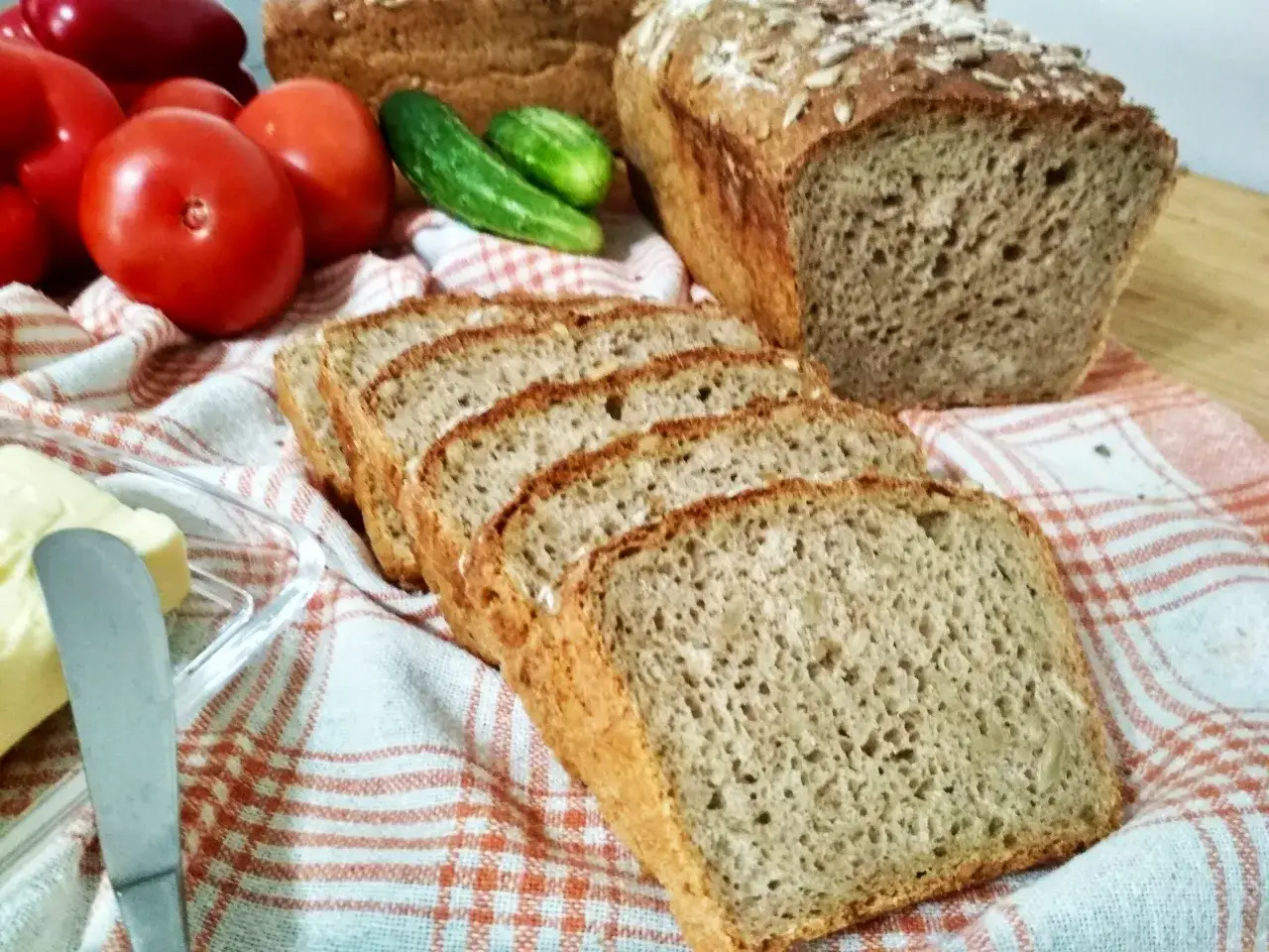 Pyszny chleb słonecznikowy pokrojony w kromki, obok pomidory, ogórki i masło. Idealny na śniadanie.