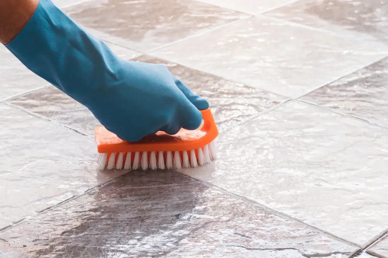 Mit Hausmitteln die Farbe vom Boden entfernen: Eine Hand in blauer Gummihandschuh schrubbt mit orangefarbener B&uuml;rste &uuml;ber Fliesen.
