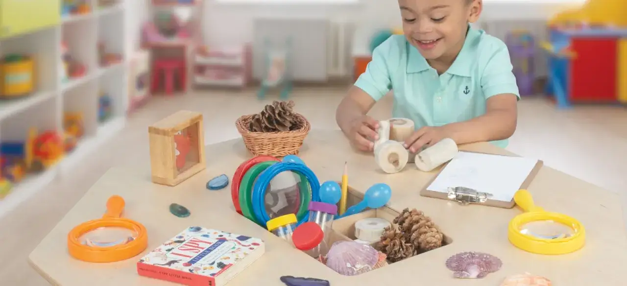 Ein Junge erkundet in der Lernwerkstatt Kindergarten Materialien wie Zapfen und Muscheln. Lupen und ein Buch laden zum Entdecken ein.