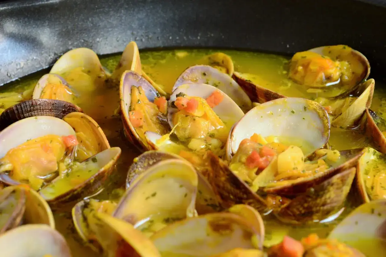 Almejas con jam&oacute;n en una sart&eacute;n, con trocitos de pimiento rojo y perejil picado en una salsa dorada.