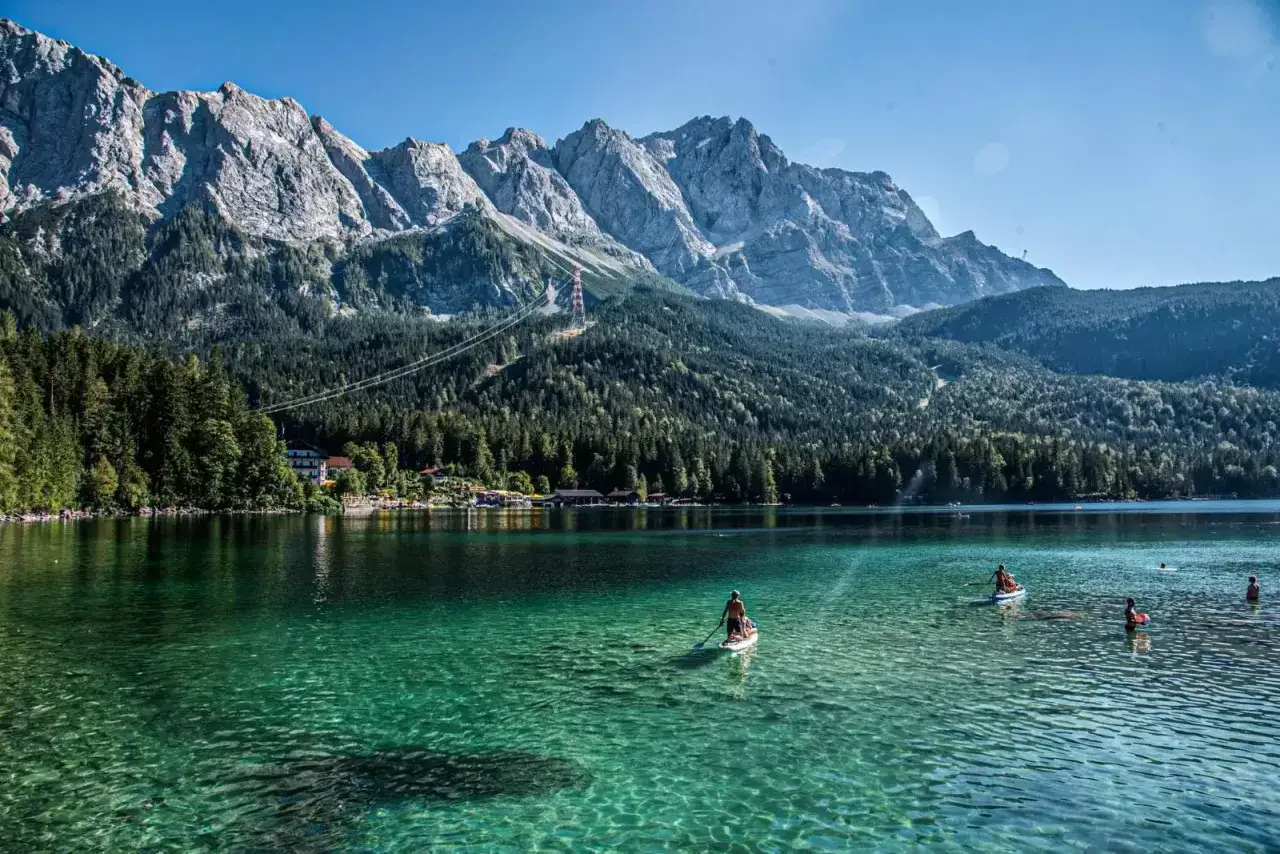 Ein Mann auf einem SUP-Board genießt den kristallklaren, blauen See in Bayern, umgeben von majestätischen Bergen und grünen Wäldern.