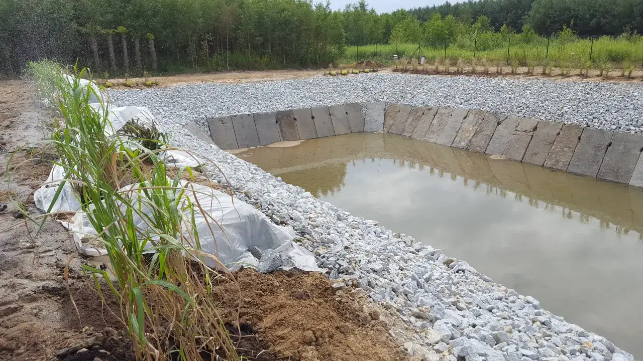 Trwa budowa stawu rybnego. Betonowe płyty i kamienie tworzą brzegi, a woda już wypełnia część niecki.