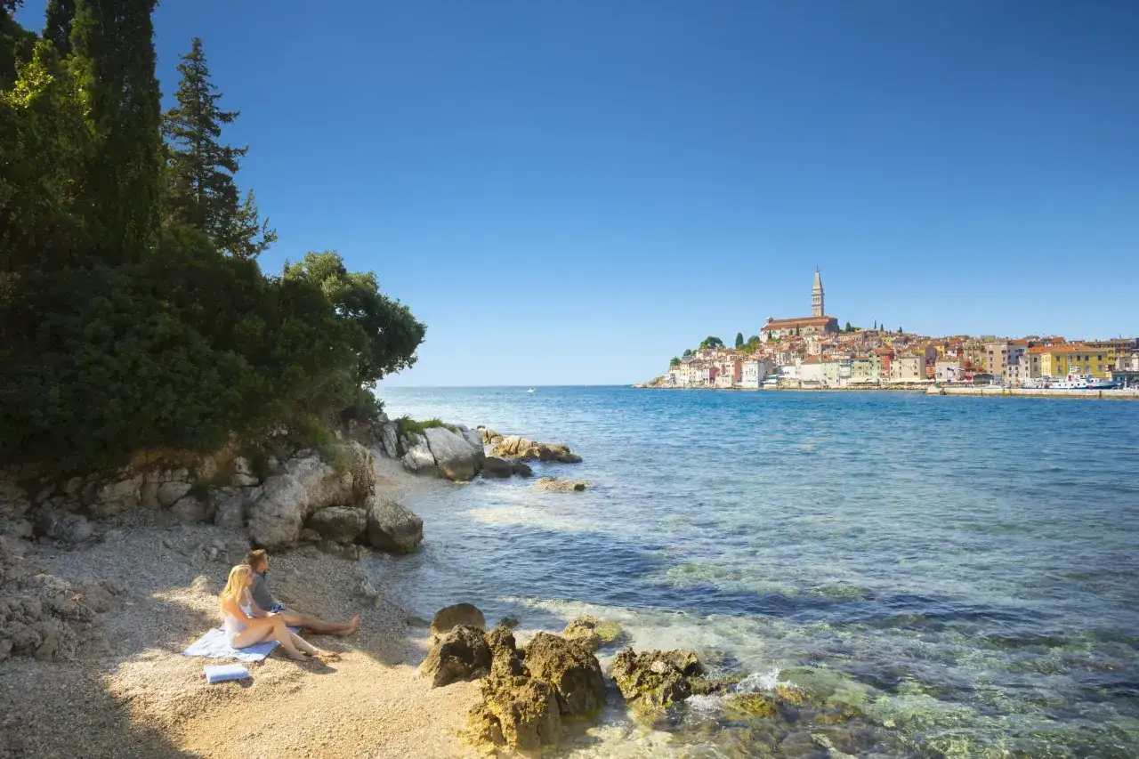 Para relaksuje się na plaży w Rovinj, Chorwacja, z widokiem na urokliwe miasto i turkusowe morze.