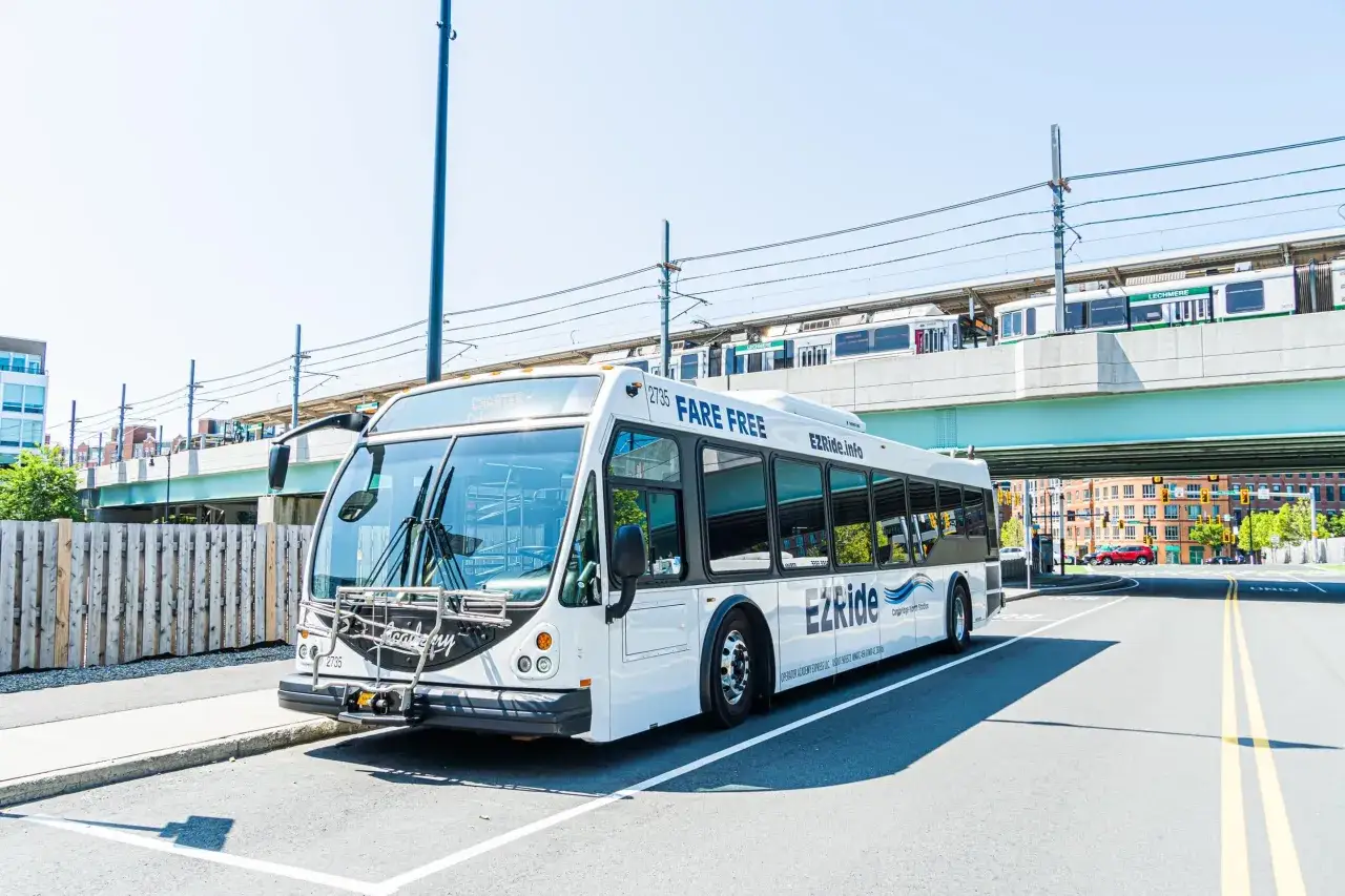A white EZRide bus, fare-free for travel from Boston to Cambridge, MA, drives past a train station on a sunny day.