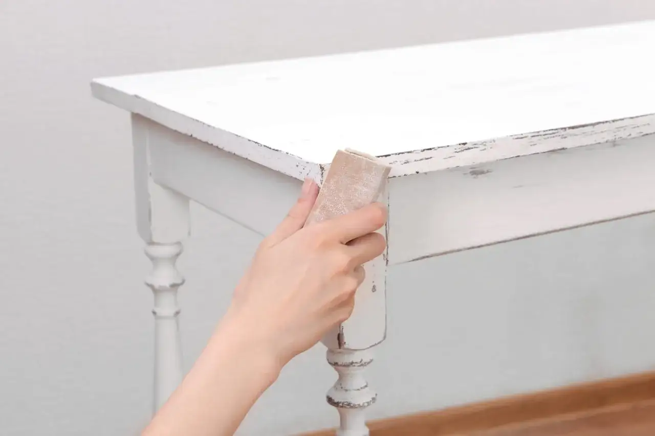 Main d'une personne utilisant du papier de verre pour décaper peinture sur bois d'une table blanche vieillie.