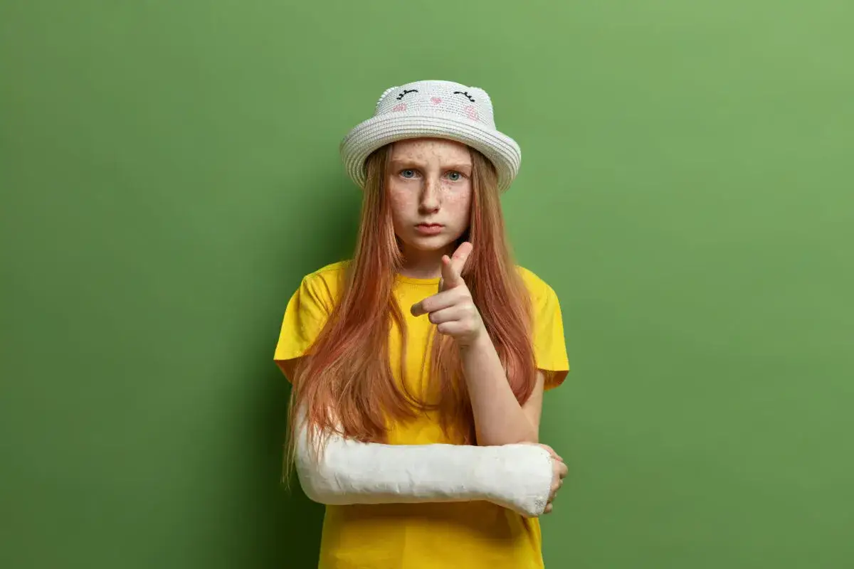 Dziewczynka z gipsem na ręce i kapeluszem na głowie pokazuje palcem, jakby wyrażała swoje zachowania nastolatków w okresie dojrzewania.