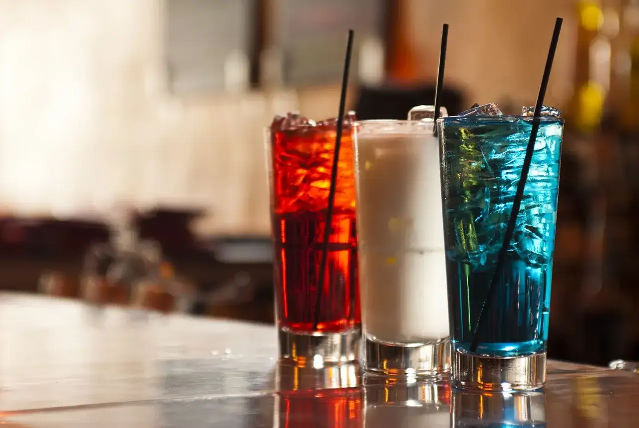 Drei bunte Longdrinks mit Eis und Strohhalmen auf einer Bar. Eine rote, eine weiße und eine blaue Variante, die Lust auf die nächste Longdrinks Liste machen.