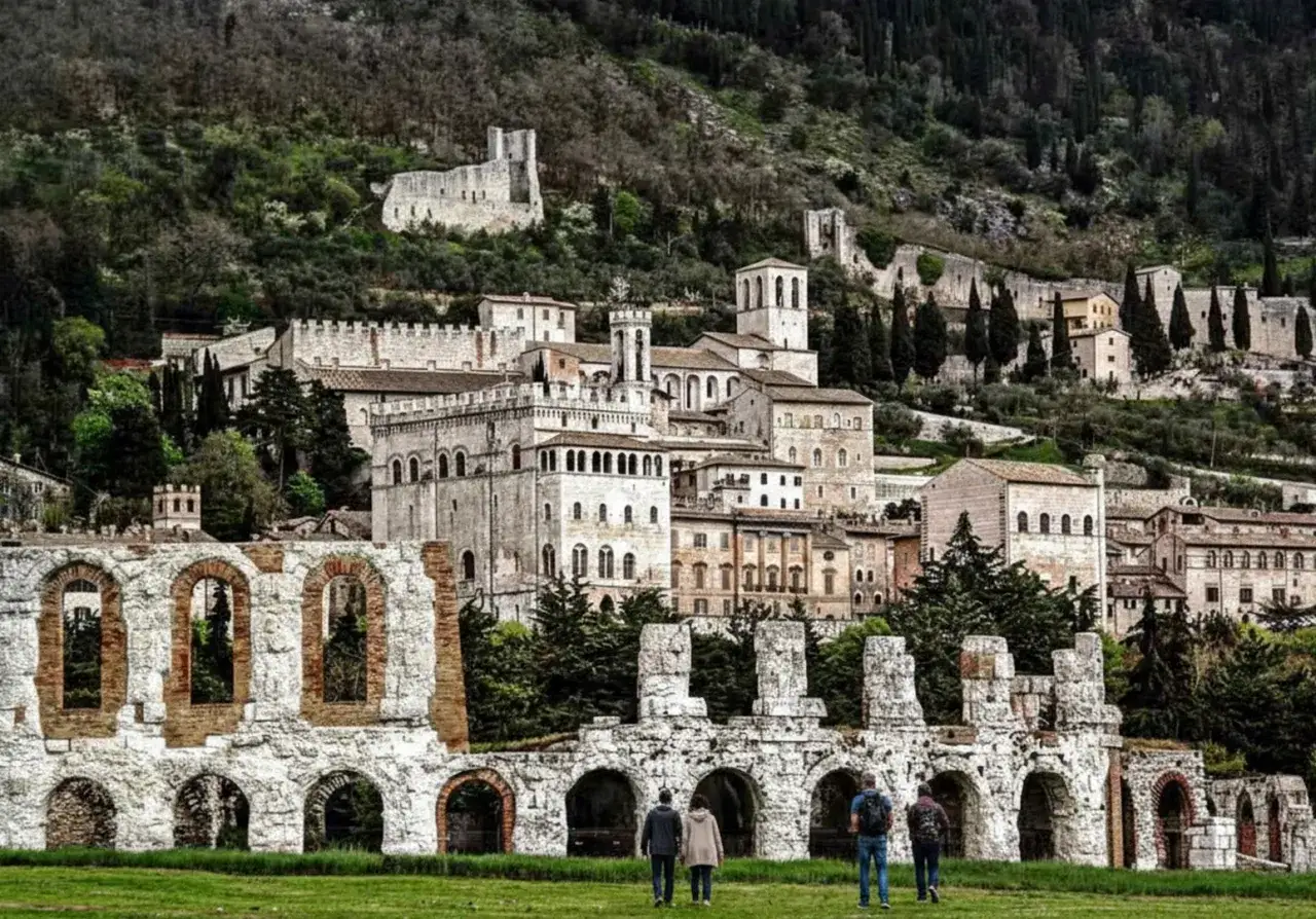 Antiche rovine romane e il centro storico medievale di Gubbio si stagliano su un colle verdeggiante.