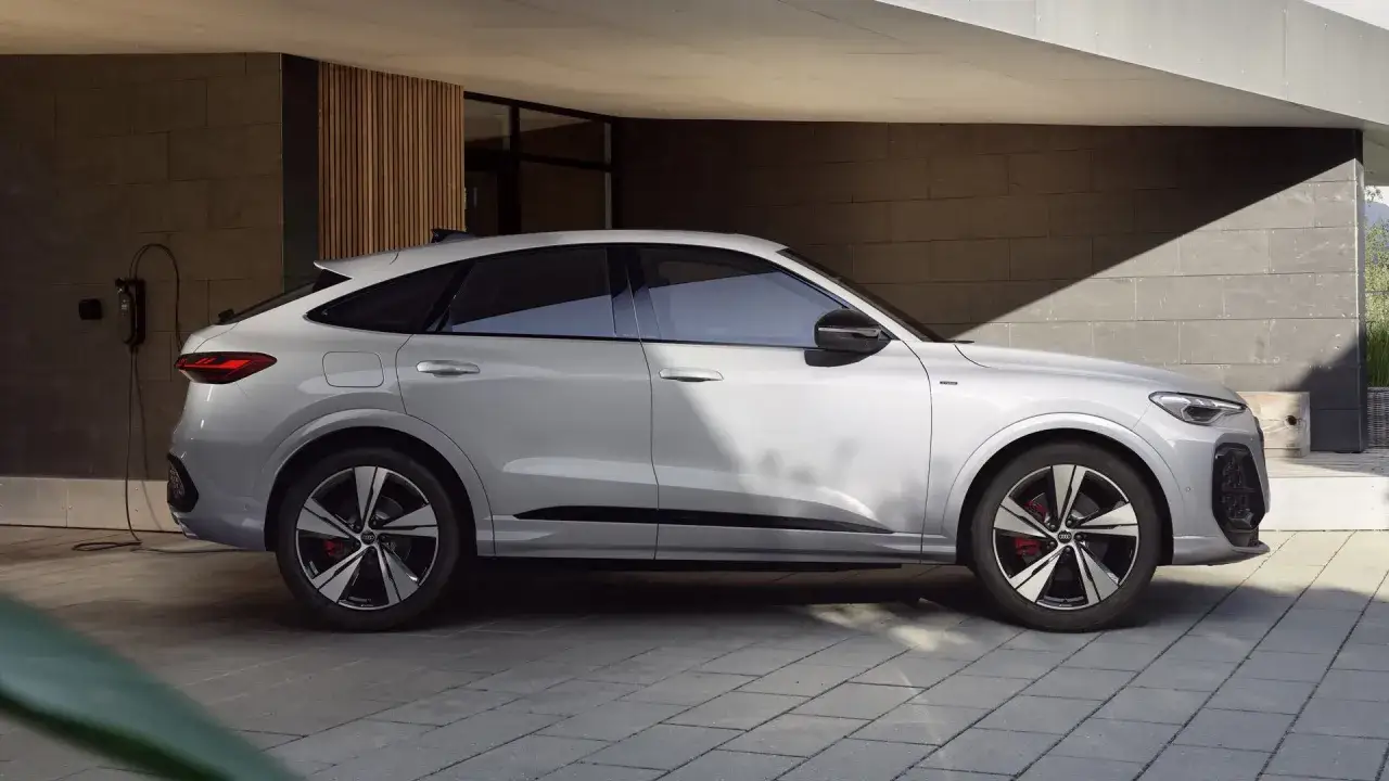 Srebrny Audi Q5 Sportback 2025 ładowany przy nowoczesnym domu. Elegancka linia nadwozia i sportowe felgi.