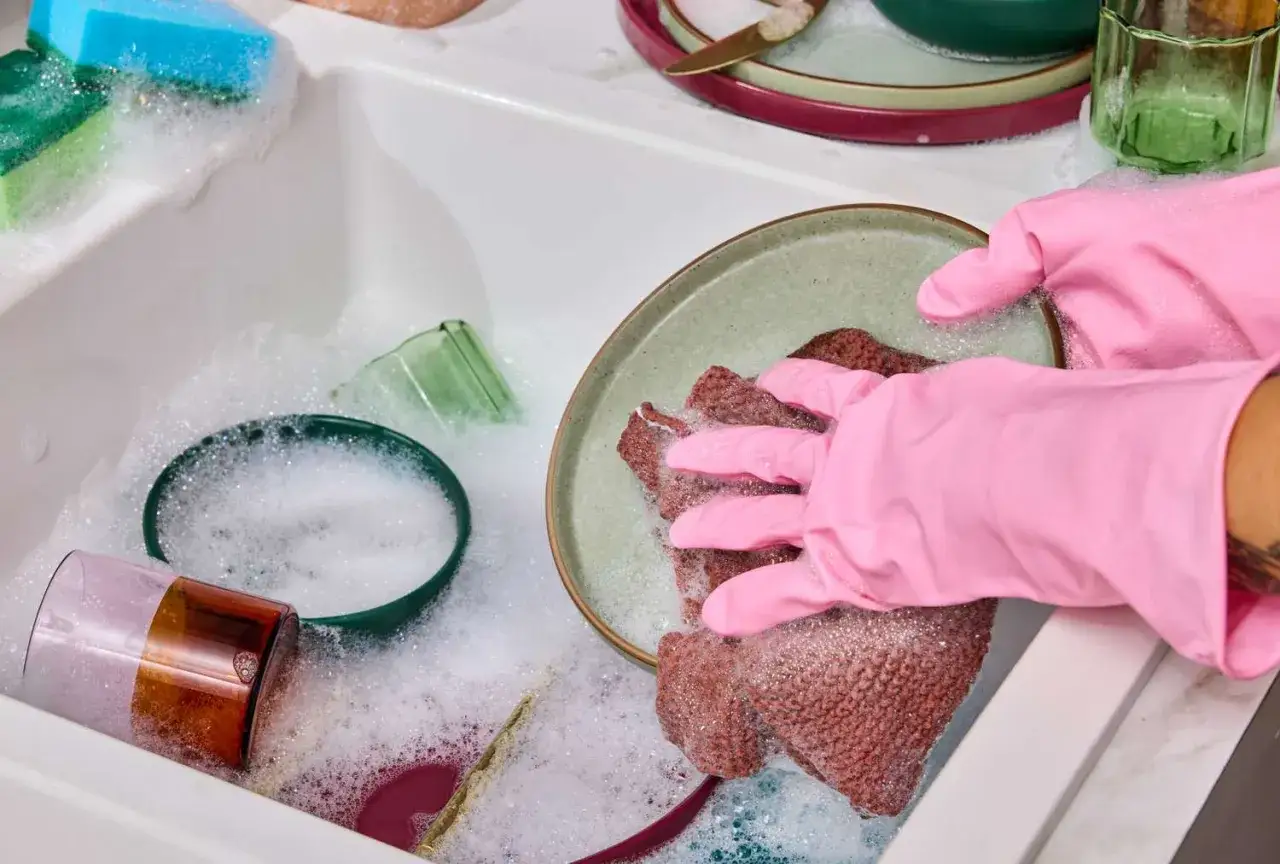 H&auml;nde in rosa Handschuhen schrubben mit einem braunen Schwamm ein Geschirr in einem seifigen Sp&uuml;lbecken.