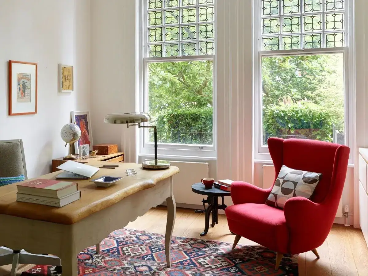 Aménagement bureau petit espace : bureau en bois, fauteuil rouge, tapis coloré, lampes, livres et fenêtres lumineuses.