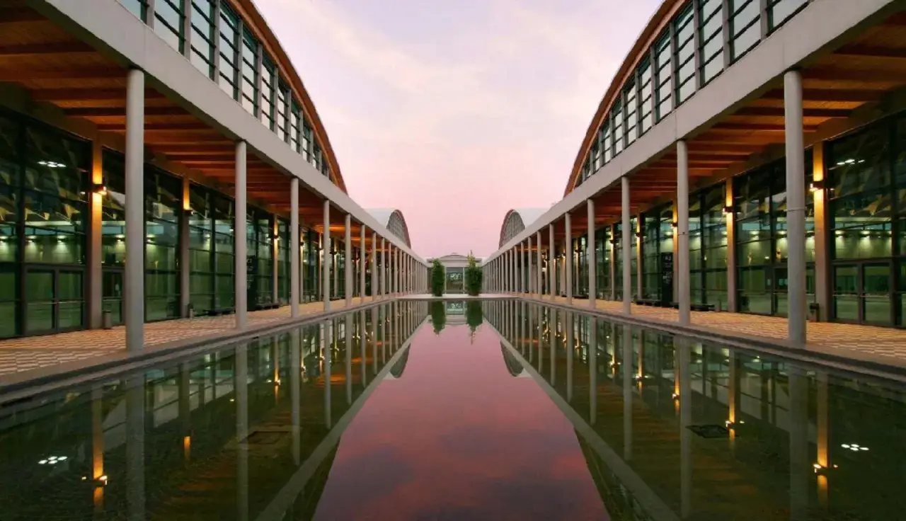 Ingresso moderno della fiera di Rimini, con specchio d'acqua e cielo rosa al tramonto. Ideale per eventi in ottobre.
