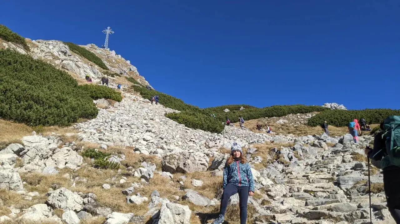 Dziewczyna na szlaku na Giewont, w tle widać krzyż i ludzi wspinających się po kamienistym zboczu.