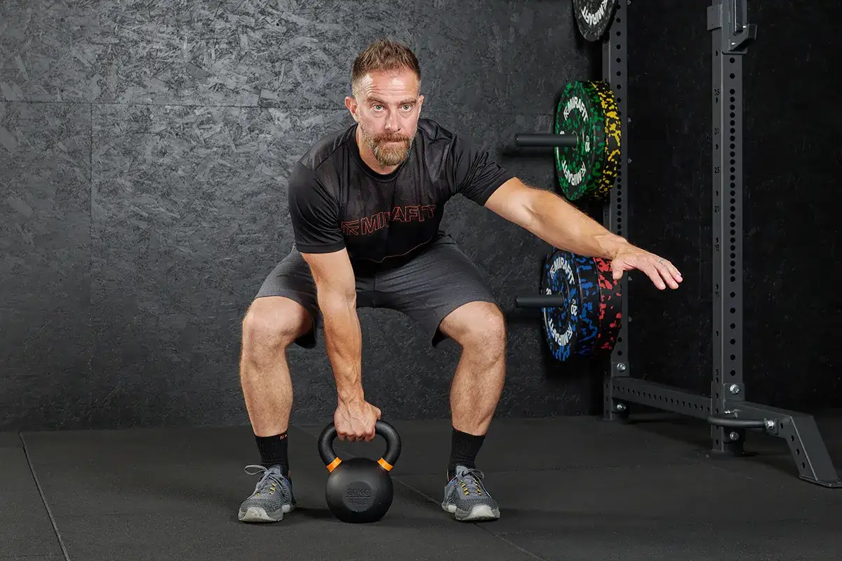 Mann mit Kettlebell in tiefer Hocke, bereit für sein **hit trainingsplan**. Gewichte im Rack.