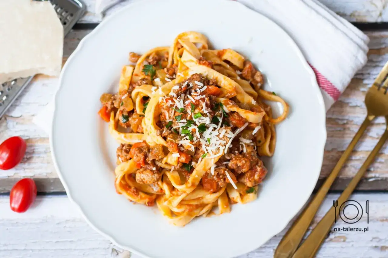 Pyszne spaghetti bolognese z tagliatelle, posypane parmezanem i natką pietruszki, na białym talerzu.