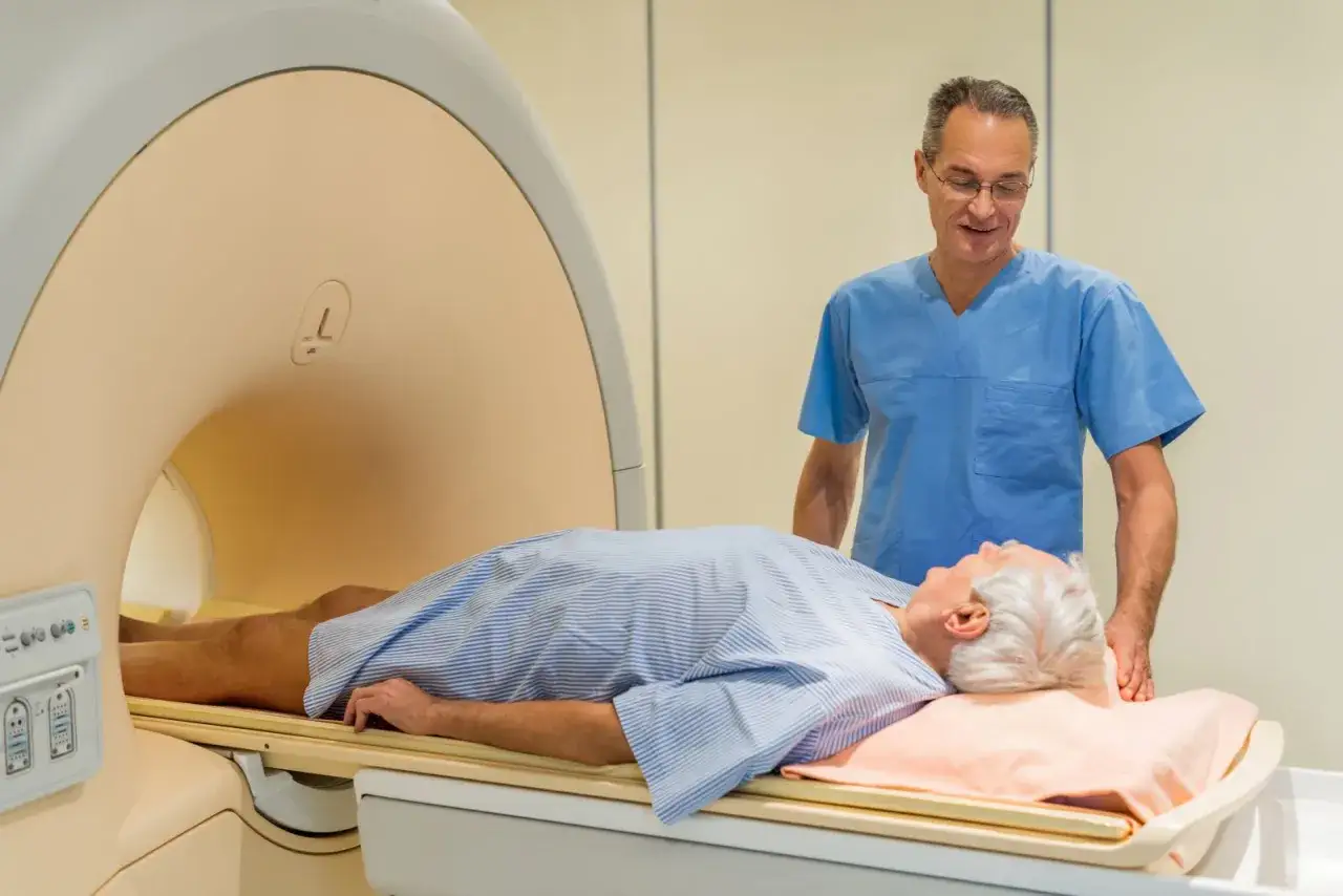 Starszy mężczyzna leży na stole do rezonansu magnetycznego, przygotowywany do badania. Lekarz w niebieskim fartuchu czuwa nad przebiegiem rezonansu magnetycznego prostaty.