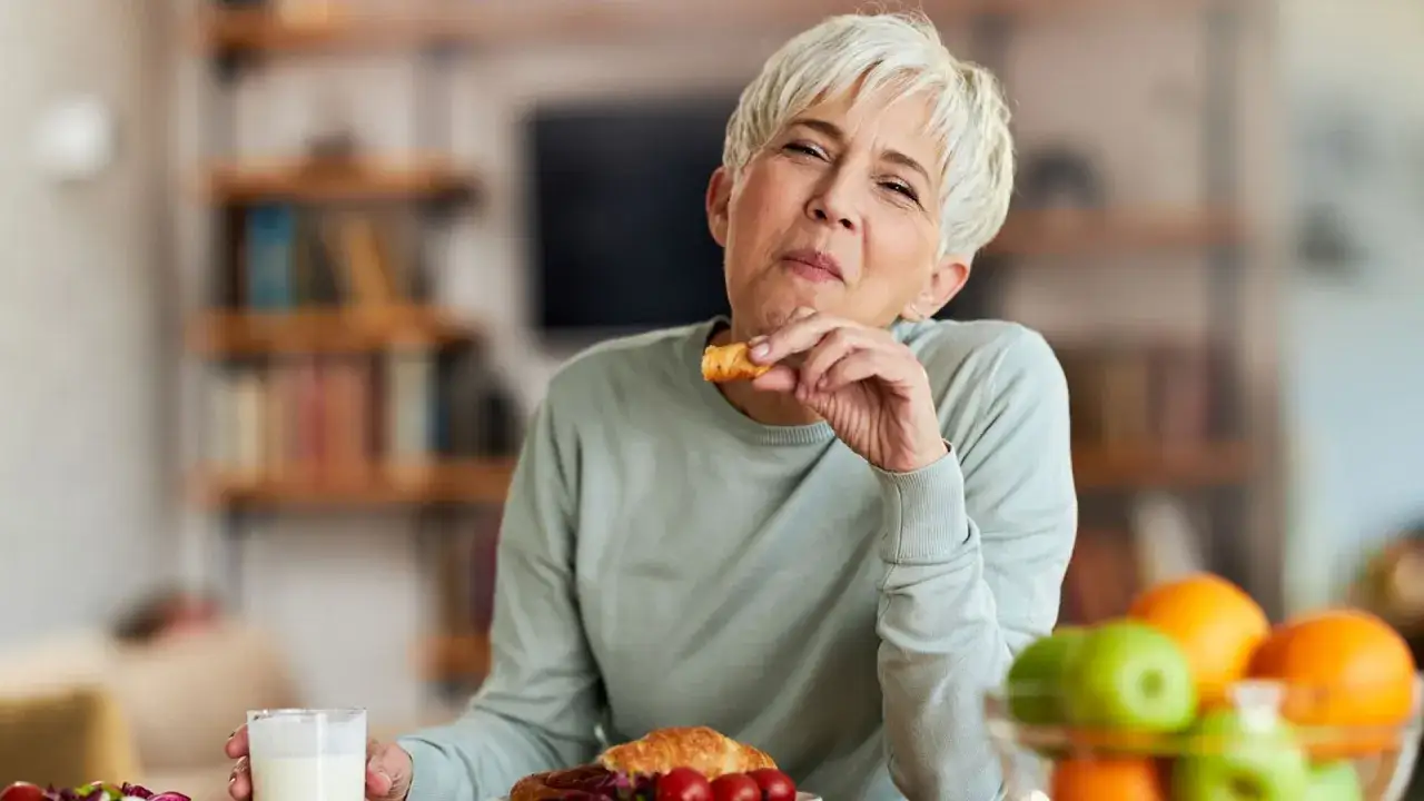 Mujer mayor comiendo un croissant, reflexionando si la menopausia adelgaza o engorda. En la mesa hay frutas y un vaso de leche.