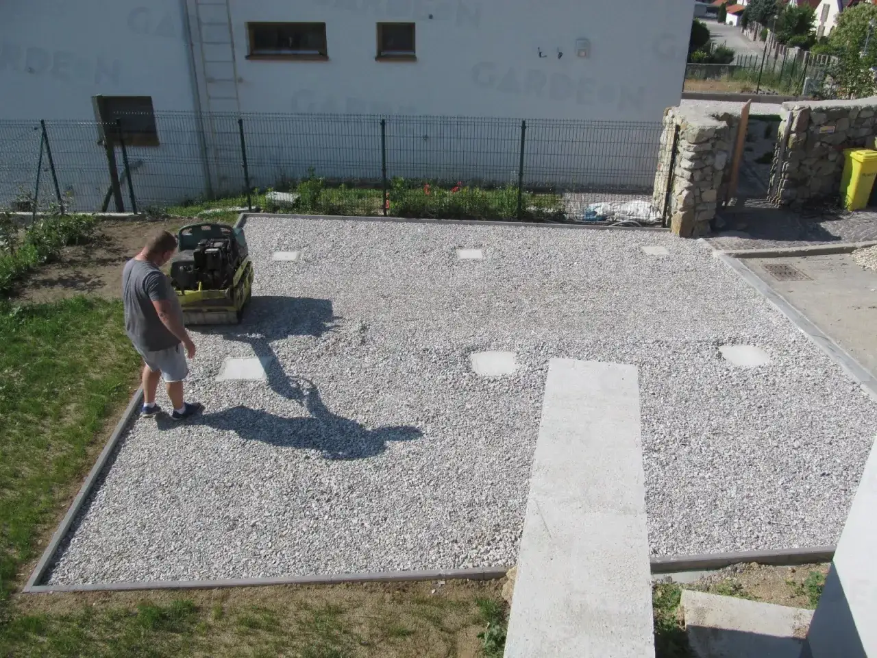 Mann verdichtet Schotter f&uuml;r einen neuen **Fundament beton**. Kleine Betonplatten sind bereits im Kiesbett eingelassen.